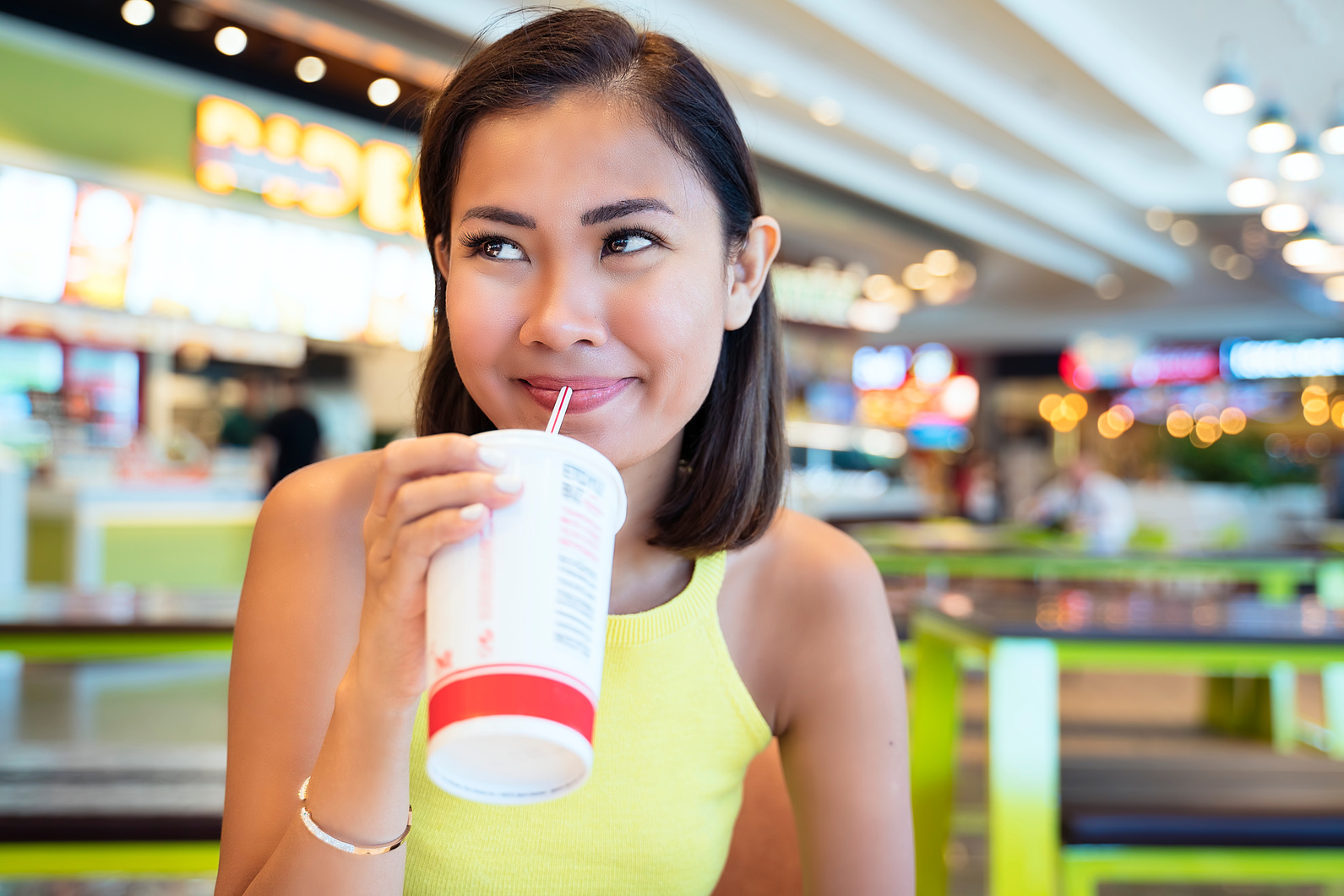 Asian woman having fast food, drinking with disposable cup in shopping mall food court